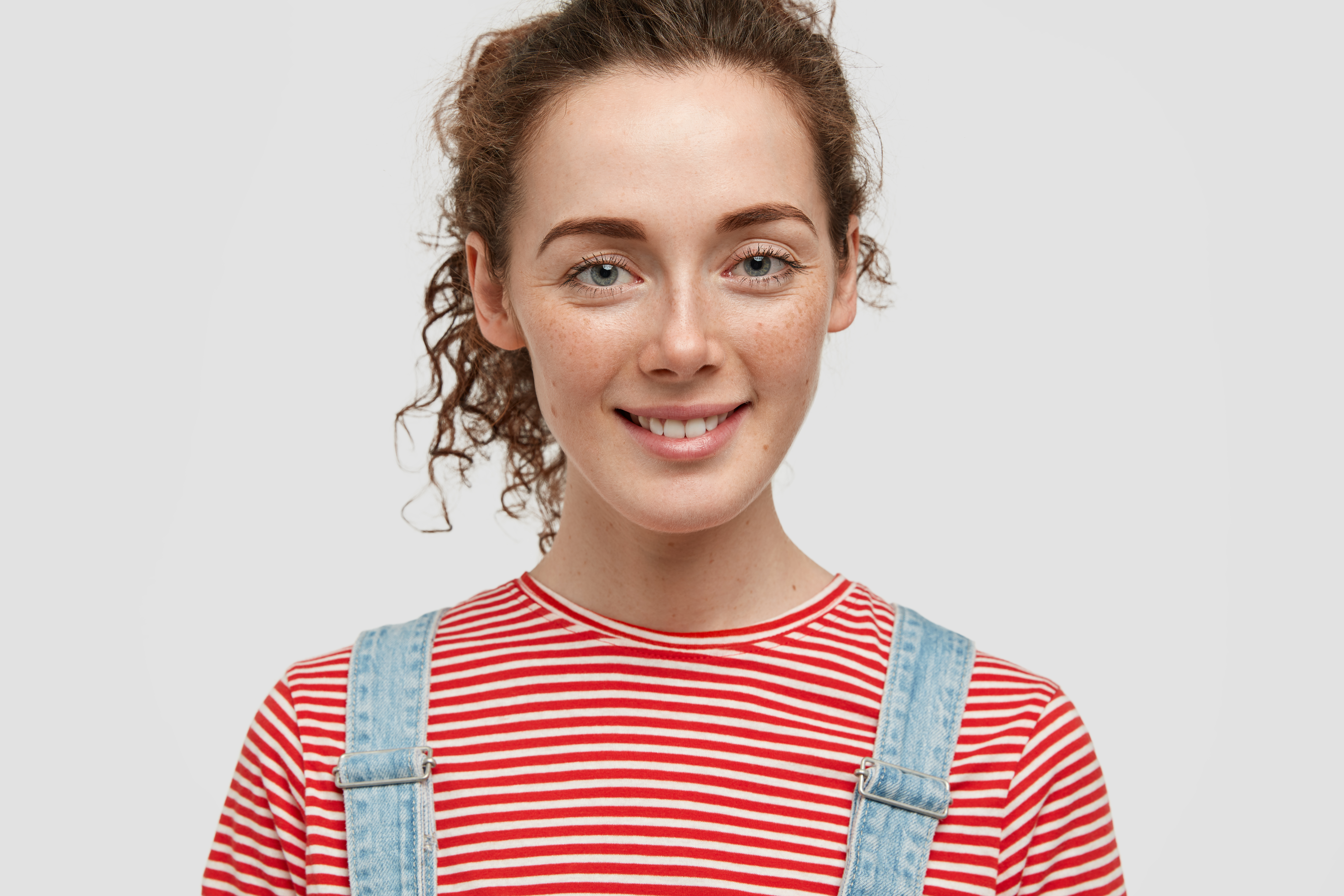 Close up shot of attractive freckled woman with curly hair, smiles positively, being in good mood after successful day, has healthy freckled skin, dressed in striped t shirt and denim overalls