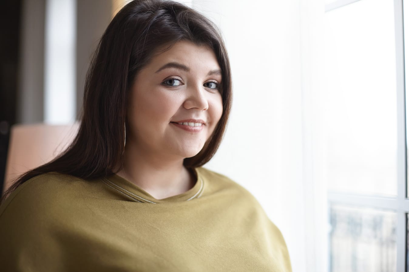 Indoor image of charming elegant young twenty year old female with chubby cheeks in stylish sweatshirt and earrings, having joyful look, posing against window background with copy space for your text